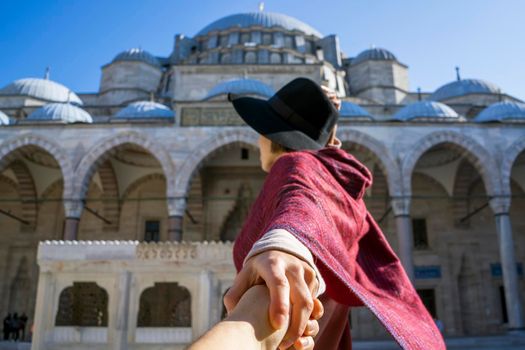 A young traveler girl in a red poncho and hat holds a friend's hand, follow me gesture. A couple in love travels to Arab countries. An ancient blue mosque on the background, Istanbul, Turkey.