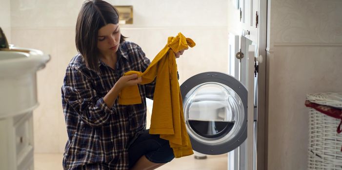 A young girl puts things in the washing machine, a woman sits on the floor of a cozy house and looks at the stain on her jacket, which needs to be removed with detergent.