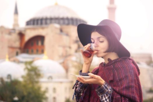 A young girl in a burgundy poncho and a hat holds traditional Turkish tea in hands. Hagia Sophia mosque in the rays of the sunset on the background. Travel in Turkey, Istanbul
