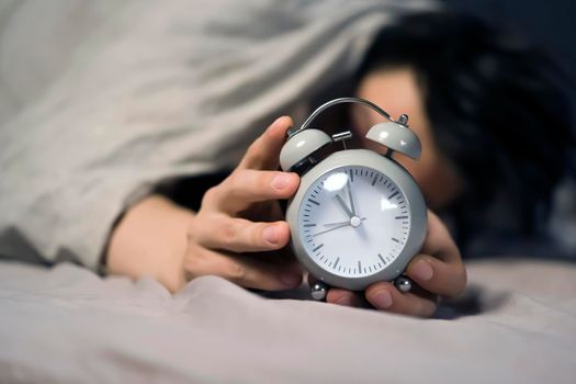 Hands of a young man from under the blankets hold a retro vintage alarm clock in gray. The person holds a clock, need to wake up.