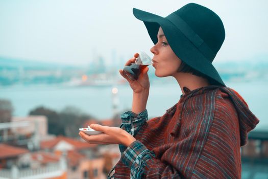 A young girl in a poncho and a hat drinks fragrant delicious tea on the background of the ocean while traveling.