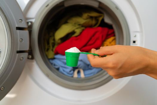 A woman's hand holds a cap with washing powder against the background of a washing machine with bright things put into it. The girl washes and wringes out things in the laundry room at home.