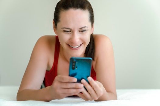 Young woman communicates via a smartphone video camera with smile on her face lying on bed at home.