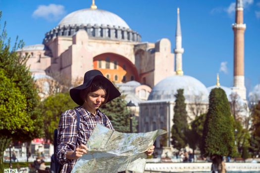 A young girl in a hat looks at a map of Istanbul on a sunny day. Beautiful ancient Hagia Sophia Mosque on the background. Traveling to Arab countries.