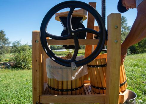 Beautiful old fashioned hand crank cider press with grinder and ground apples ready for pressing. Agricultural nature background with green grass and farm fields, shot in natural light with one man and copy space.