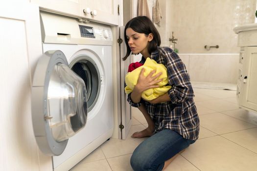 A young girl puts things in the washing machine, a woman sits on the floor of a cozy house and looks at clothes that need to be cleaned with washing powder.