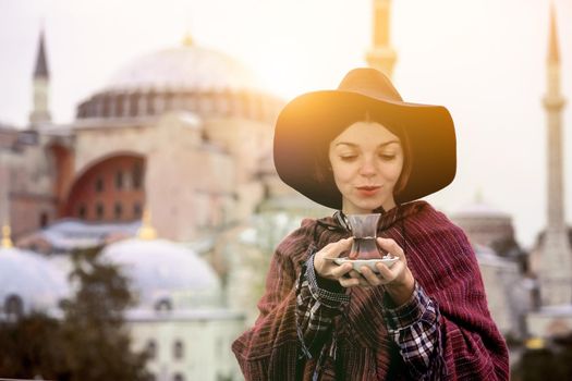 A young girl in a burgundy poncho and a hat holds traditional Turkish tea in hands. Hagia Sophia mosque in the rays of the sunset on the background. Travel in Turkey, Istanbul