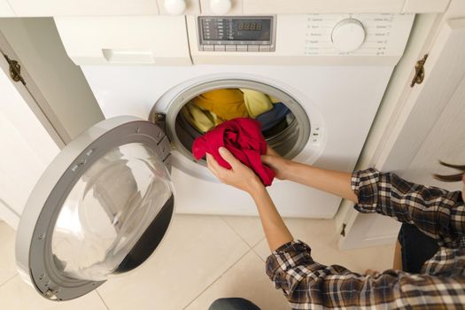 A girl folds bright clothes into a new washing machine in the bathroom at home, a woman washes things and spin them out with detergent in the laundry close-up, top view.