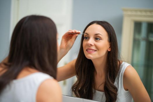 Young woman is plucking eyebrows in bathroom.