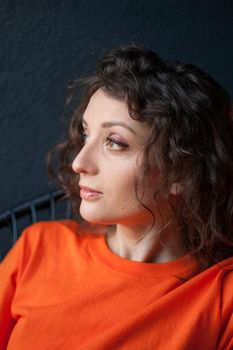 Female portrait of a beautiful curly girl in bright orange t-shirt at home in her apartment on dark background, happy people concept.