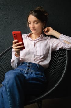 Beautiful curly girl is using red smartphone at home in her apartment, on dark background, happy people concept.