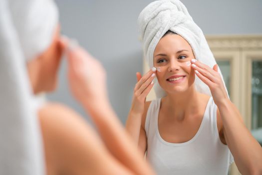 Portrait of young woman who is applying skin cream on her face.