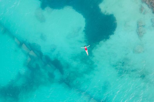 Aerial top view of woman wearing red swimsuit lying on a transparent turquoise water surface on caribbean beach. Travel and vacations concept. Tropical background with copy space