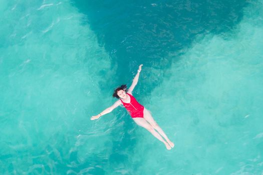 Aerial top view of woman wearing red swimsuit lying on a transparent turquoise water surface on caribbean beach. Travel and vacations concept. Tropical background with copy space