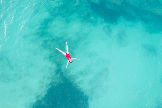 Aerial top view of woman wearing red swimsuit lying on a transparent turquoise water surface on caribbean beach. Travel and vacations concept. Tropical background with copy space