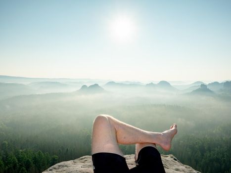 View above barefoot male body in black pants sleeping on cliff sticking out from morning fog.  Natural park in Sachsen, Deutschland