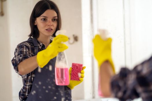 A young girl is cleaning the bathroom, applying detergent with a spray and washing the mirror with a sponge in yellow gloves on her hands. Smiling woman taking care of the cleanliness of her home.