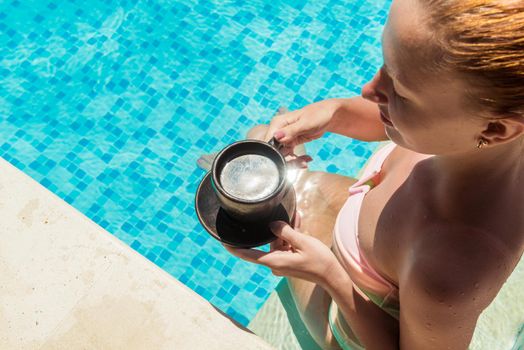 Young woman wearing swimsuit enjoying coffee near swimming pool at exotic location