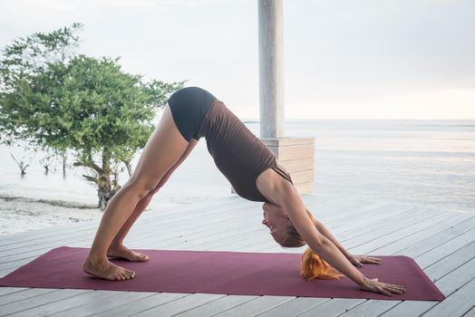 Young slim lady doing yoga downward facing dog on a beach with sunset over Bali in the background