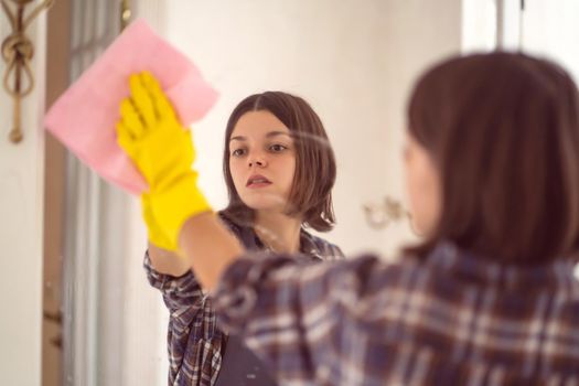 A young girl is cleaning the bathroom, applying detergent with a spray and washing the mirror with a sponge in yellow gloves on her hands. Smiling woman taking care of the cleanliness of her home.