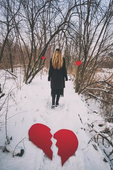 Brokenhearted woman walking away from broken red heart in forest in winter.