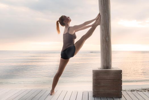 Young slim lady doing yoga stretching exercises on a beach with sunset over Bali in the background