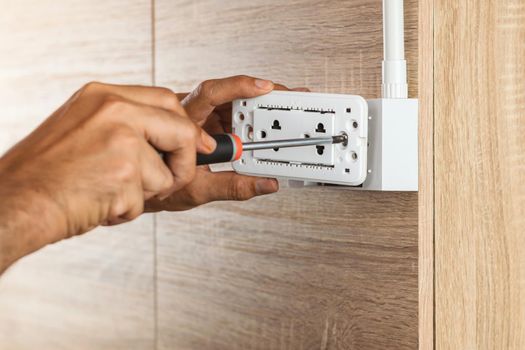 Electrician is using a screwdriver to install the electric power socket in to a plastic outlet box on a wooden wall.