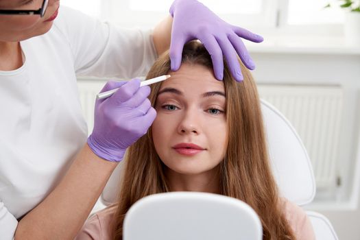 Cosmetologist using facial marker on patient face before beauty treatment in spa salon. Client is looking into mirror in in beauty clinic