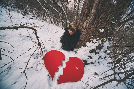Depressed and brokenhearted woman leaning on tree in forest in winter.