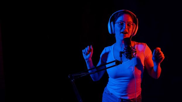 Caucasian woman in glasses and headphones sings into a microphone in neon light on a black background. An emotional girl is recording a song in a recording studio.