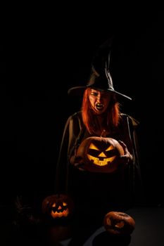 An evil witch holds a halloween jack-o-lantern glowing in the dark.