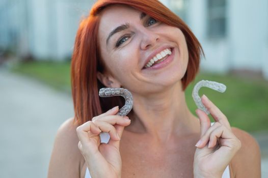 Red-haired Caucasian woman holding transparent mouthguards for bite correction outdoors. A girl with a beautiful snow-white smile uses silicone braces.