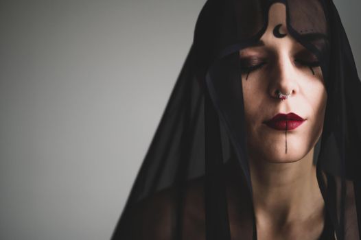 Redhead woman with a black veil on her head in a dark studio. A girl with an earring in her nose smiles slyly. Witch Makeup. Halloween costume