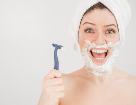 Cheerful caucasian woman with a towel on her head and shaving foam on her face holds a razor on a white background.
