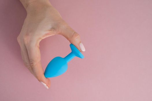 A woman is holding a blue anal plug on a pink background. Adult toy for alternative sex.