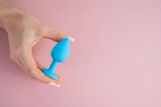 A woman is holding a blue butt plug on a pink background. Adult toy for alternative sex.