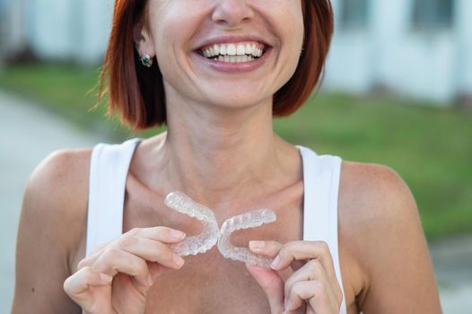 Red-haired Caucasian woman holding transparent mouthguards for bite correction outdoors. A girl with a beautiful snow-white smile uses silicone braces.