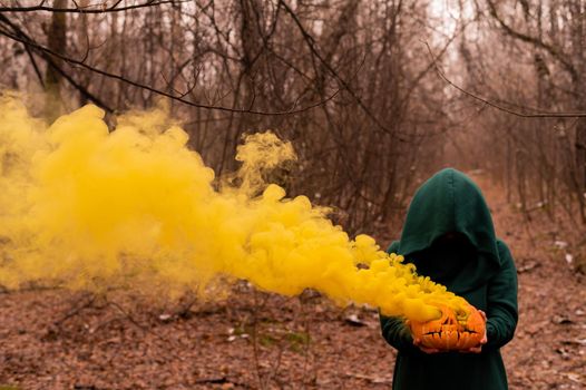 A creepy witch holds a steaming pumpkin in a deep forest. Jack o lantern emits yellow smoke for halloween.