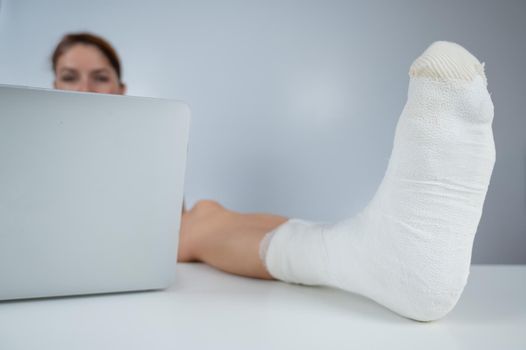 Caucasian woman lifted her leg with plaster to work desk and works on laptop on white background.