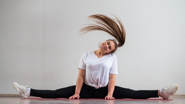 A young flexible fat woman sits in a transverse twine and waves her hair against a white background.