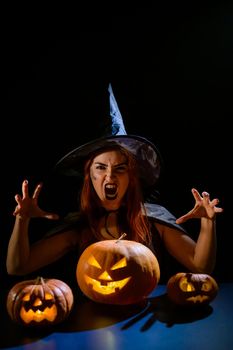 An ominous witch in a hat conjures over a jack-o-lantern. Traditional halloween characters. Mystical fog creeps over pumpkins with carved terrible faces. An evil witch casts a spell