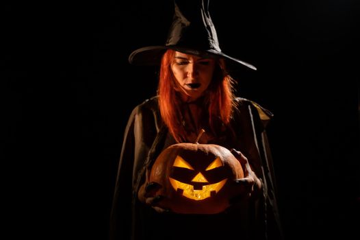 An evil witch holds a halloween jack-o-lantern glowing in the dark.