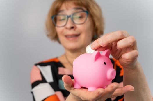 An elderly woman with glasses puts a coin in a piggy bank on a white background.