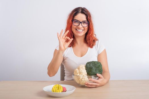 Caucasian woman prefers healthy food. Redhead girl chooses between broccoli and donuts on white background