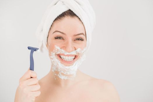 Cheerful caucasian woman with a towel on her head and shaving foam on her face holds a razor on a white background.