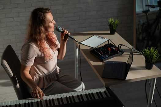 Expressive female singer has a video blog on a laptop. Portrait of a girl recording a song on a web camera and composing on a synthesizer. Online music lessons. Distance learning in quarantine