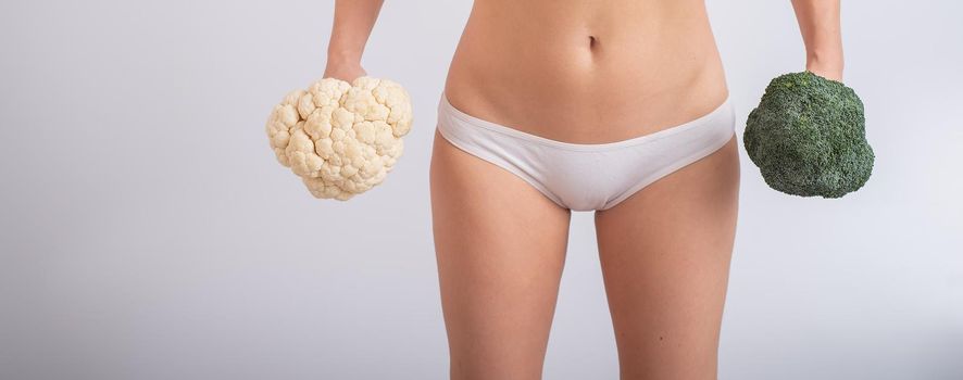 A faceless woman in panties holds cauliflower and broccoli on a white background. Food habits