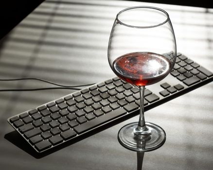 A goblet with red wine and a keyboard on a white table with shade from blinds.