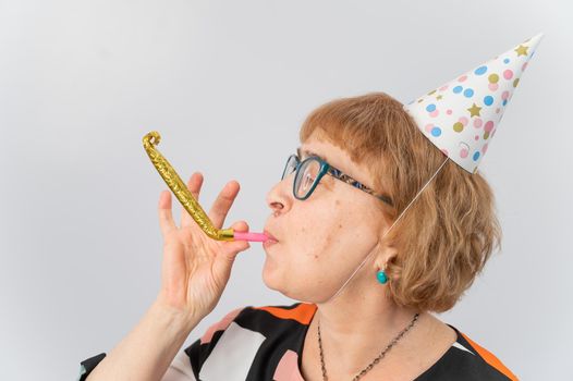 Portrait of a smiling elderly woman in a festive cap holding a whistle tongue on a white background.