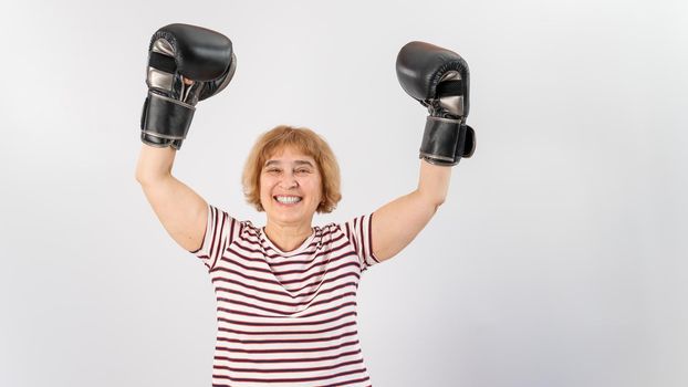 An elderly woman in fighting gloves raises her hands up on a white background. Victory in a duel.